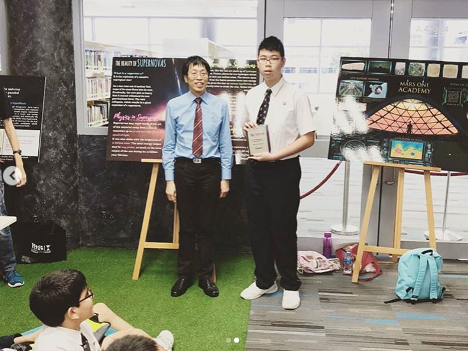 A student in a white shirt and blue tie presents at an exhibition booth, standing beside display boards with astronomy-related content and an exhibit on a table.